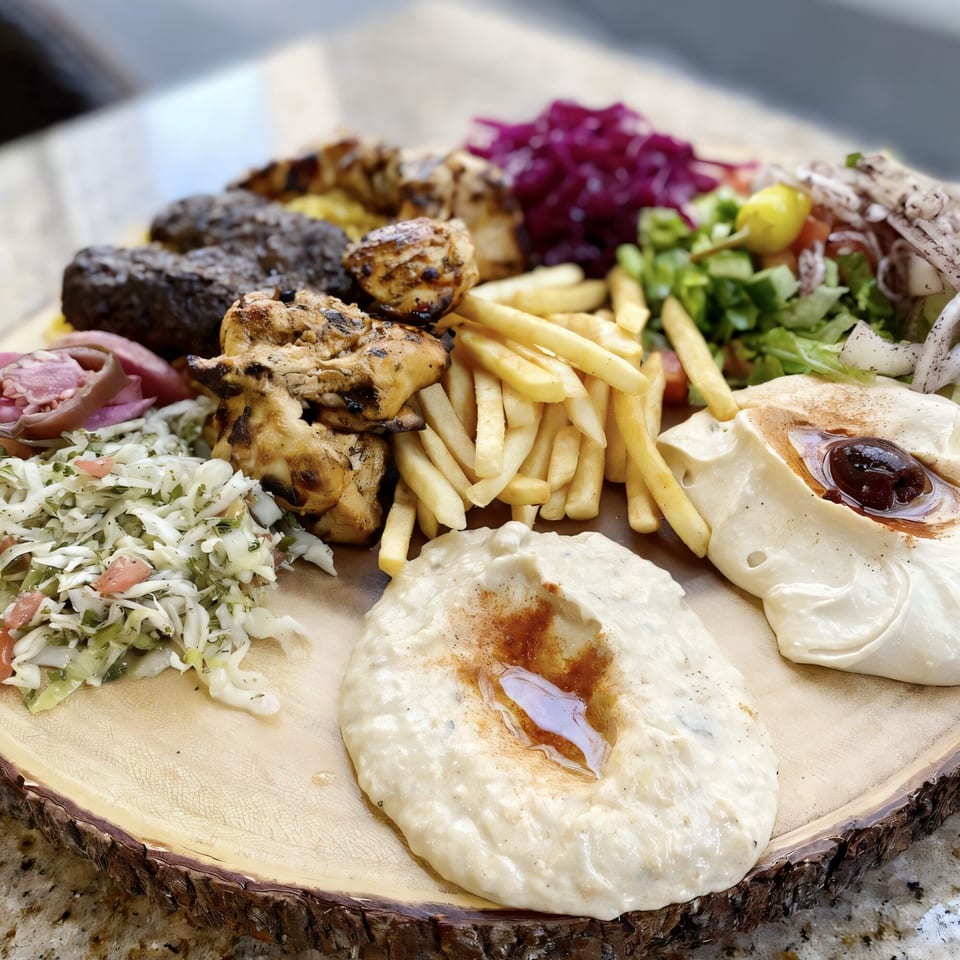 A Killer Kabab platter with grilled chicken, beef kababs, fries, salads, and hummus on a wooden board.