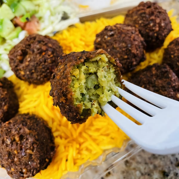 A falafel ball, broken open by a white fork, revealing its green interior, with yellow rice and salad.